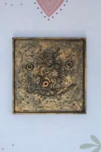 Abstract texture with metallic painting, featuring circular and gear-like shapes, intertwined lines, and beads. The tone is industrial and intricate.