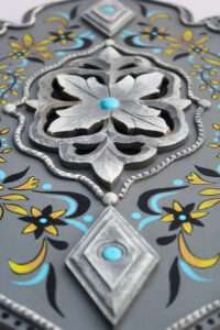 Ornate decorative panel with a central silver floral motif, accented by turquoise dots. Surrounded by yellow and black floral patterns on a gray background.