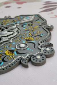 Ornate decorative panel with intricate gray, blue, and yellow floral patterns. The design features embossed silver elements and a reflective surface.