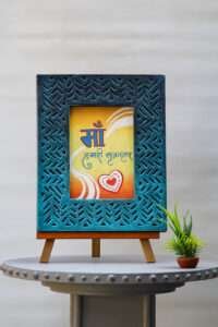 A decorative handmade wooden photo frame with intricate patterns surrounds a colorful artwork. The artwork has Hindi text, swirls, and a heart design, conveying warmth and love.