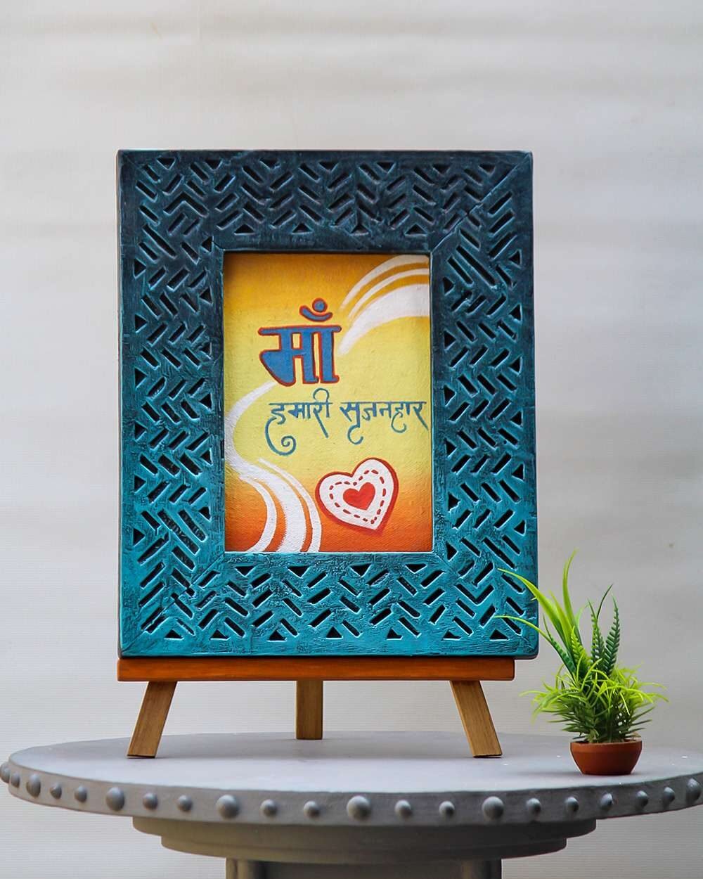 A decorative handmade wooden photo frame with intricate patterns surrounds a colorful artwork. The artwork has Hindi text, swirls, and a heart design, conveying warmth and love.