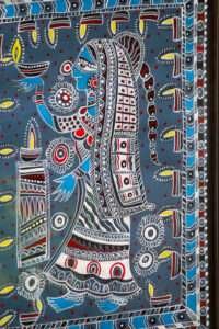 Intricate Madhubani painting depicts a woman holding a lamp, adorned in traditional attire with detailed patterns, surrounded by floral motifs on a blue background.