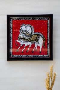 Framed painting depicting two ornately decorated white horses with intricate black and gold patterns, set against a vibrant red background with yellow dots. buy horse painting online