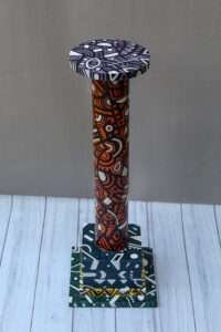 A colorful decorative side table with bold geometric and abstract patterns in orange, black, and white atop layered square bases on a wooden floor.