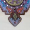 An ornate decoration features a central bronze flower surrounded by a circular pattern with red, blue, and white accents, set against a brown background.