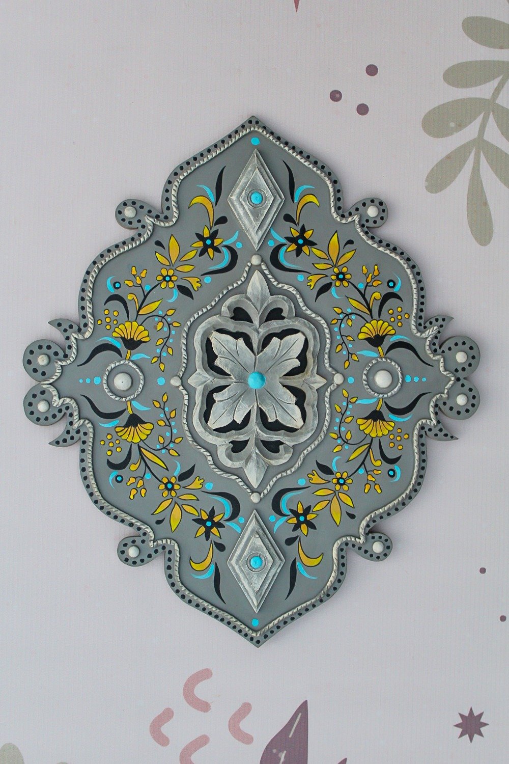 Ornate gray wall decoration with a central floral motif, surrounded by intricate yellow and blue designs. Elegant, symmetrical, and decorative.