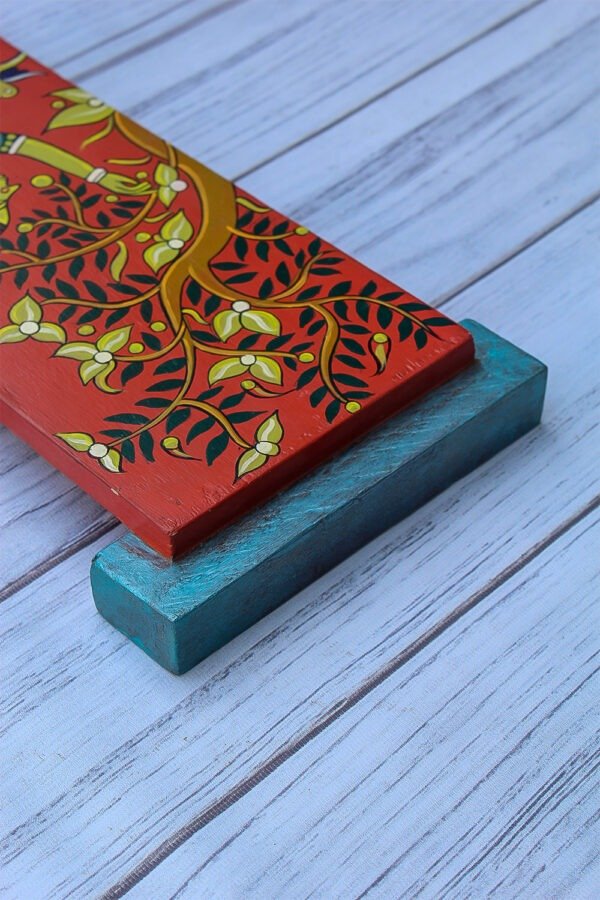 A vibrantly painted wall hanging with intricate yellow tree branches and green leaves on a red background, resting on a blue block on a light wooden floor.