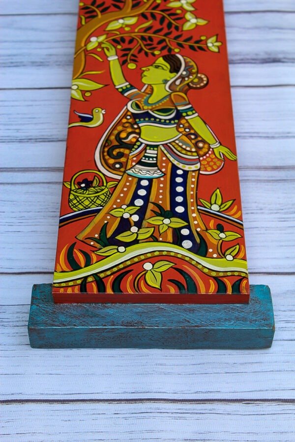 Colorful wall hanging of a woman in traditional attire reaching for a branch, with vibrant patterns and birds on a bright orange background. Set on a weathered blue stand.